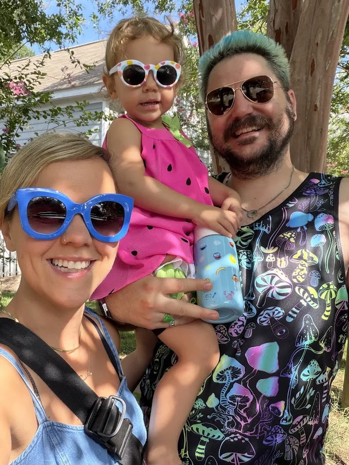 Adam, his wife and their toddler in a sunny selfie, all three in big sunglasses, Adam with teal hair and a mushroom-print tank