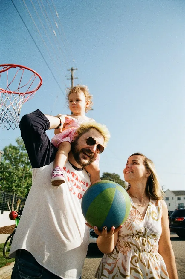 A film photo of Adam with bleached hair and his daughter on his shoulders under a basketball hoop, his wife smiling up at them with the ball