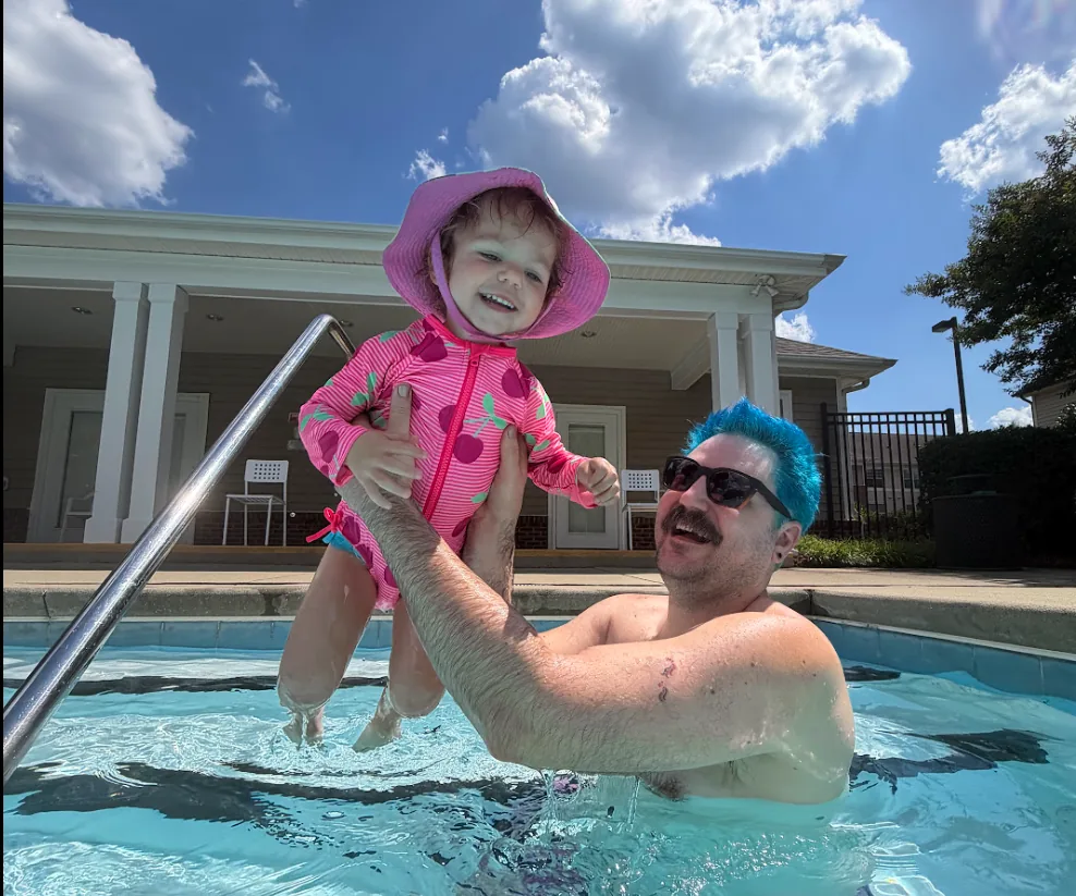 Adam with bright blue hair in a pool, lifting his laughing daughter in a pink swimsuit and sun hat