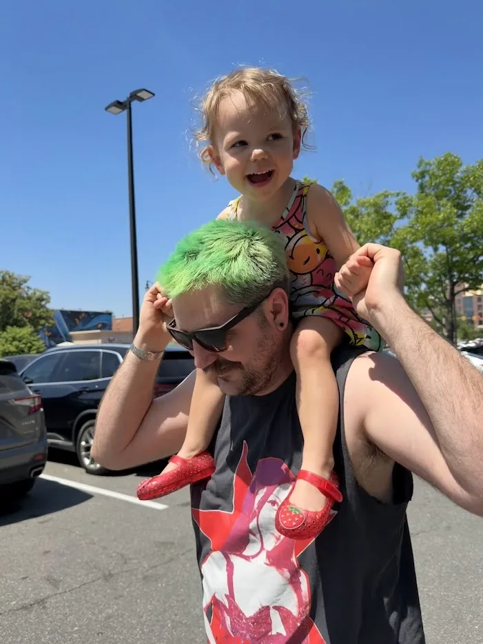Adam with bright green hair carrying his laughing daughter on his shoulders in a sunny parking lot