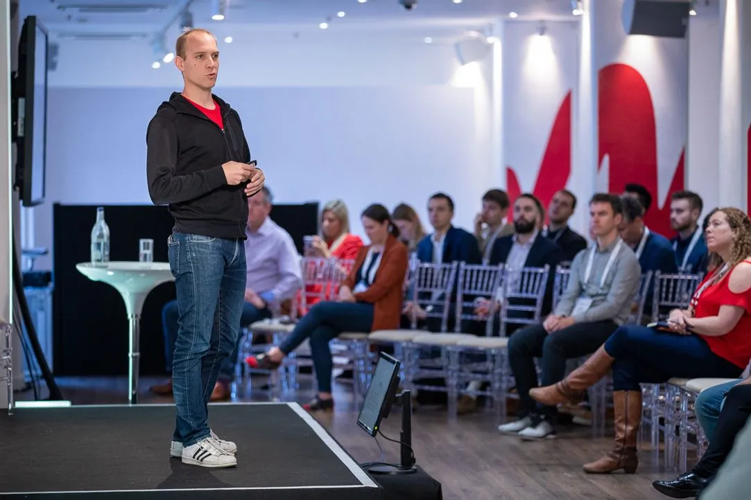 A speaker presenting to a seated audience in front of a wall painted with the red W
