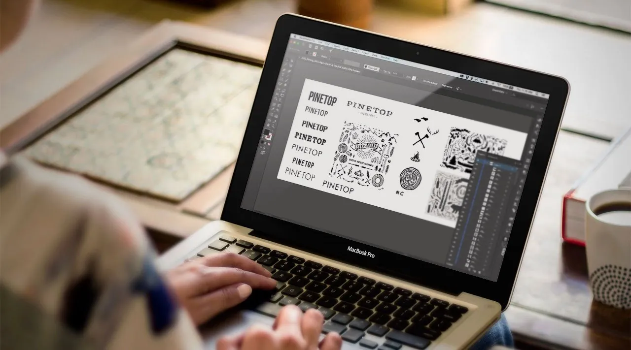 Pinetop wordmark and illustration explorations open in Illustrator on a laptop: stacked type options, axes, antlers, a campfire and a wood-ring NC badge