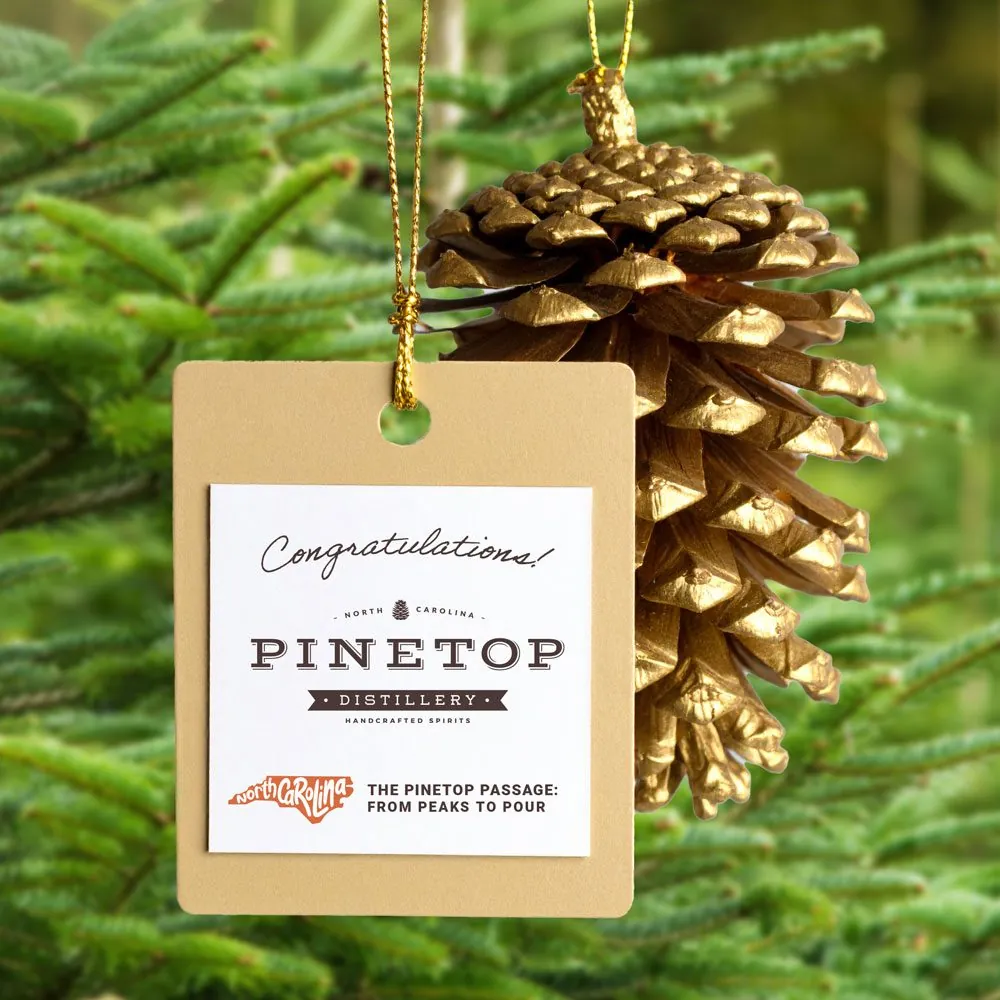 A kraft gift tag with the Pinetop Distillery lockup hanging from a gold-painted pinecone on a fir branch
