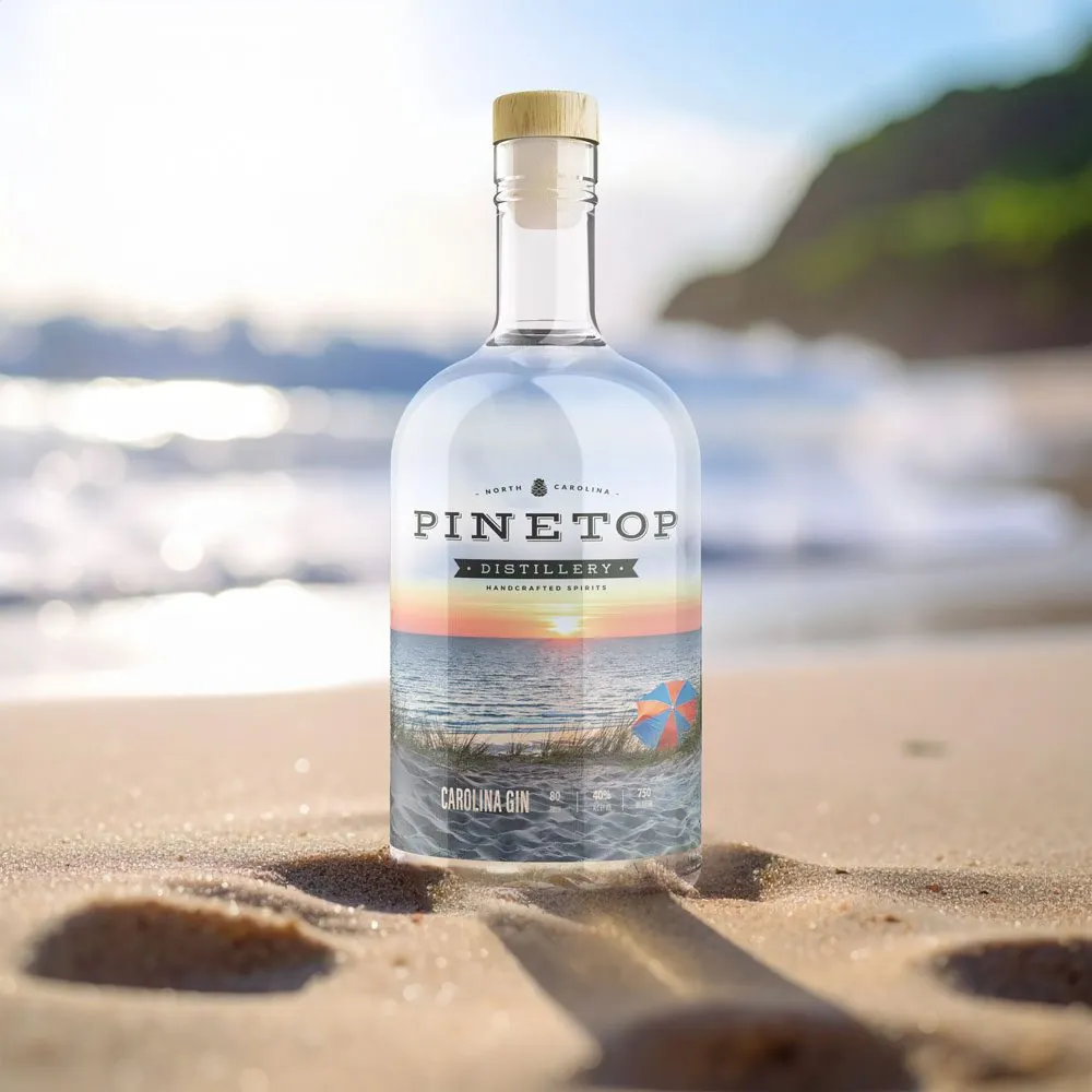 A Carolina Gin bottle standing in beach sand, its clear label showing a sunset over the ocean and a striped umbrella