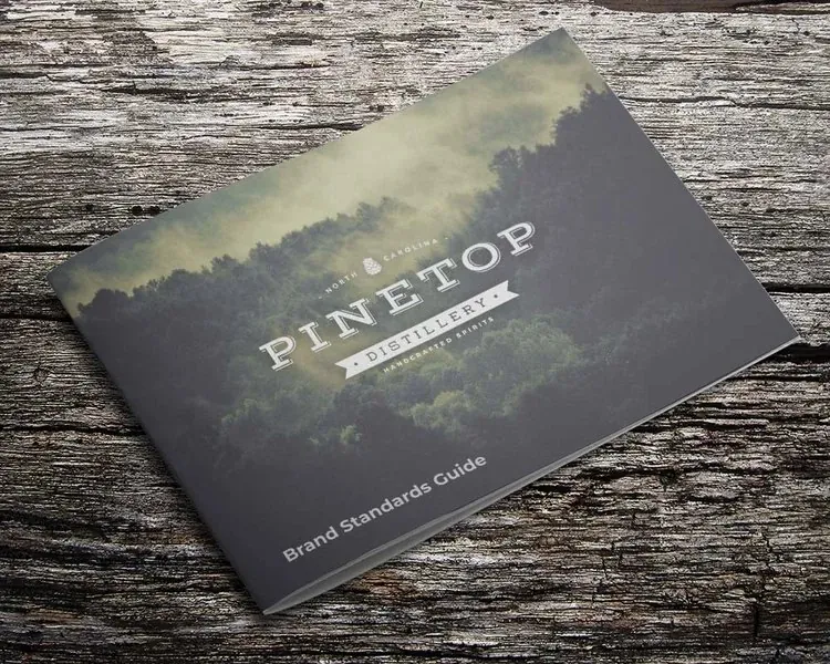 The Pinetop Distillery Brand Standards Guide on weathered wood, its cover a misty forest