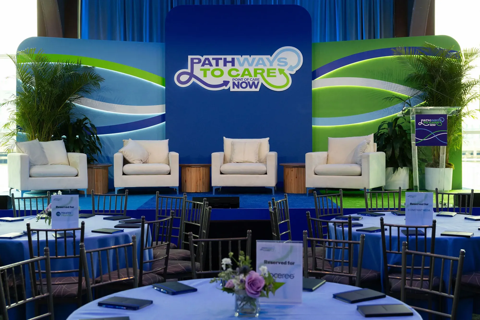 The Pathways to Care main stage at Pier Sixty: the looping logo on a deep blue wall between lit blue and green waves, four white armchairs, an acrylic podium and reserved tables in front
