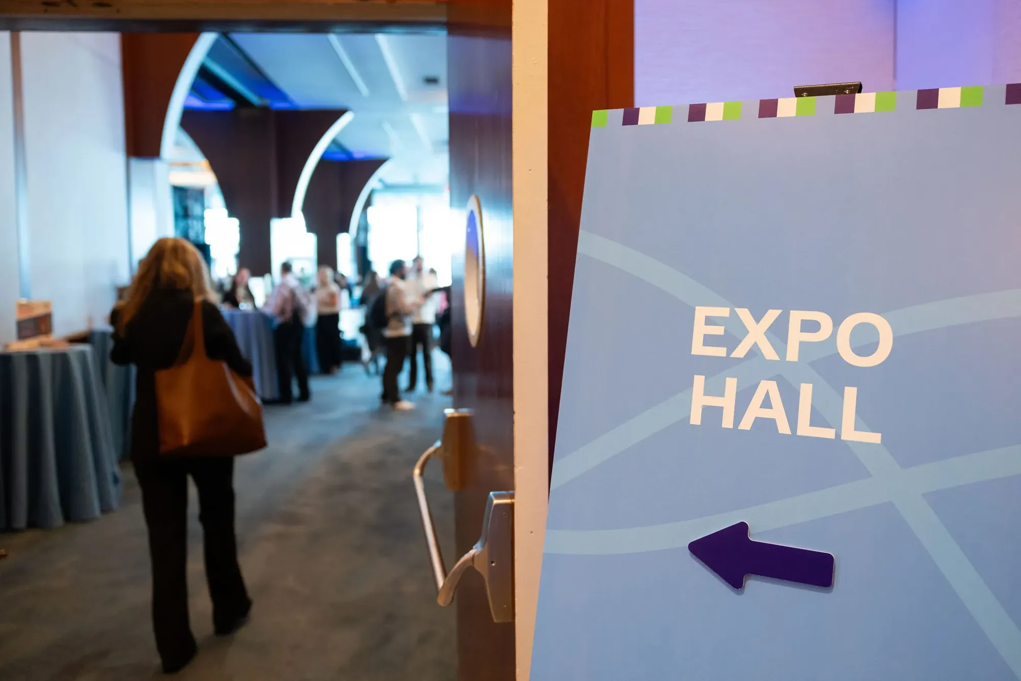 A directional sign reading Expo Hall with a purple arrow, crossing lines and a striped top edge, guests walking through the doorway beside it